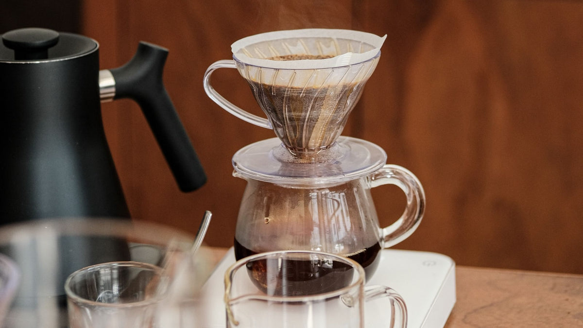 Clear pour-over dripper and glass server on a scale beside a black kettle and coffee cups