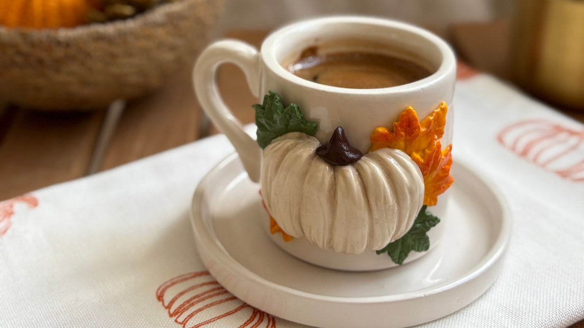 A pumpkin-decorated coffee cup on a saucer with autumn-patterned fabric