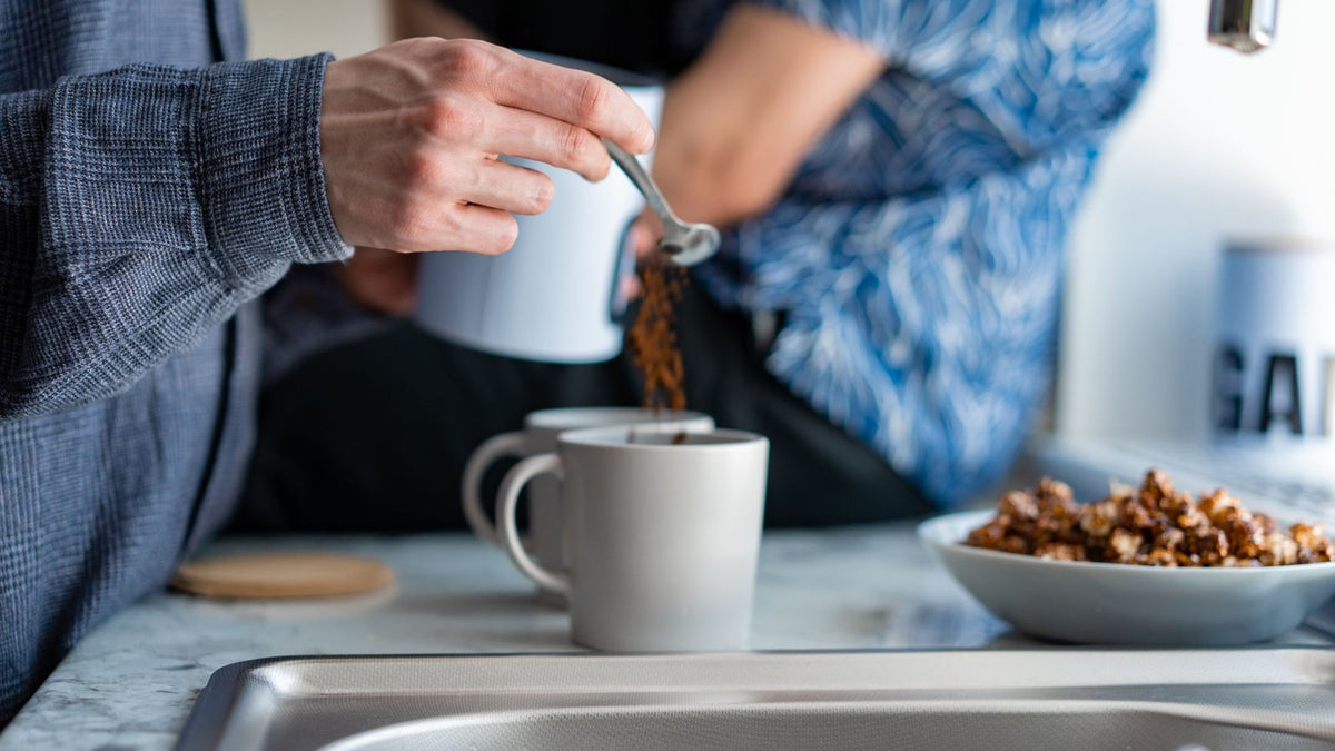 A person spooning coffee into a white mug while another person stands beside the kitchen counter