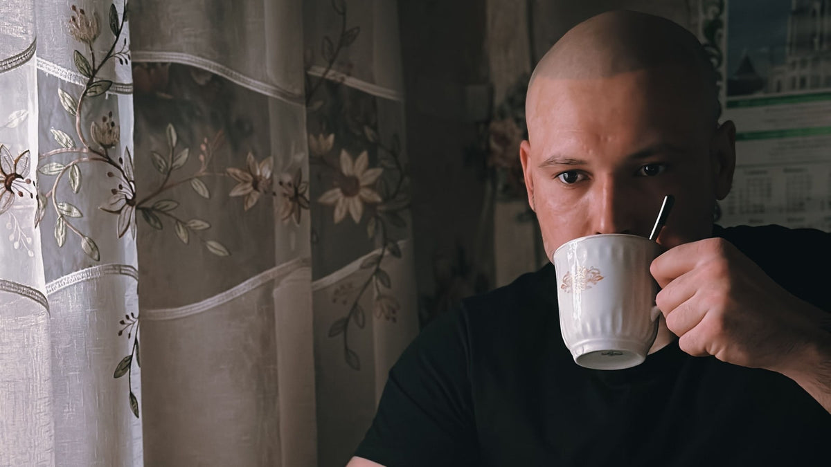 A man enjoying coffee from a ceramic mug by a curtained kitchen window
