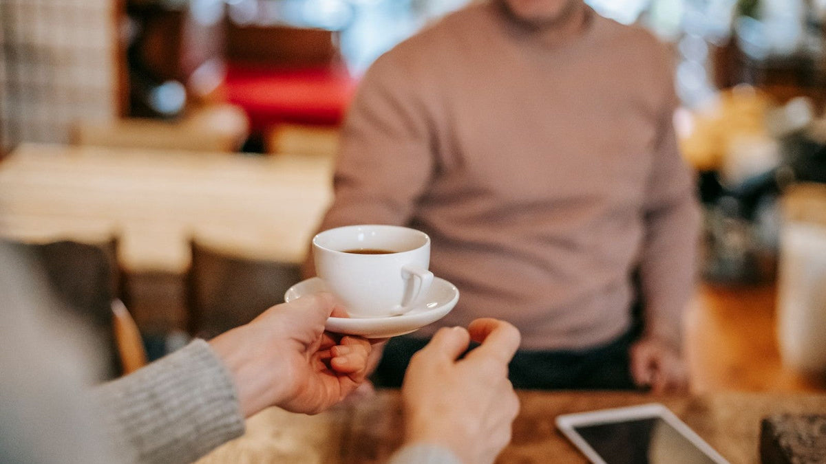One person passing a white coffee cup and saucer to another across a wooden table