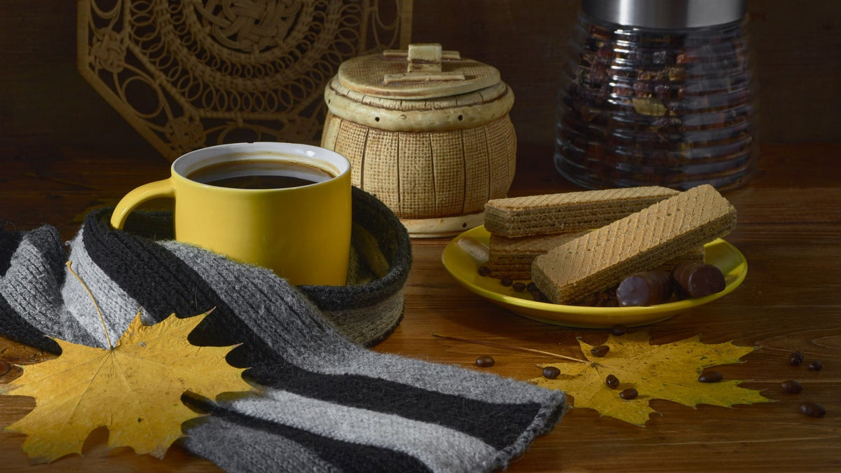 A yellow coffee cup beside wafers, a grey scarf and autumn leaves on a wooden table