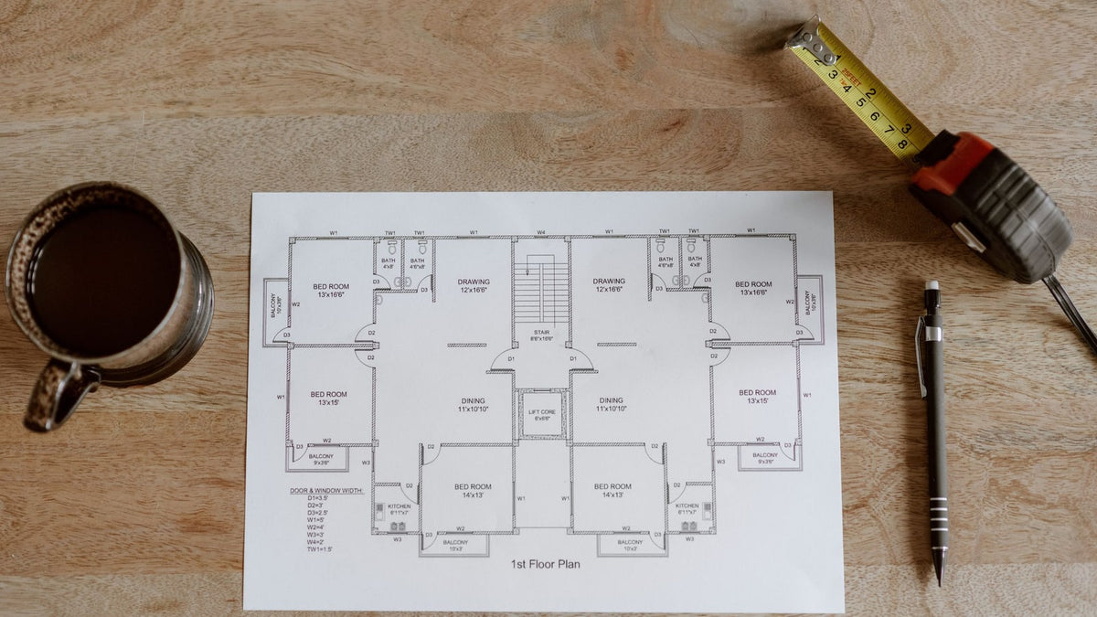 A cup of coffee, tape measure and pen beside a floor plan on a wooden table