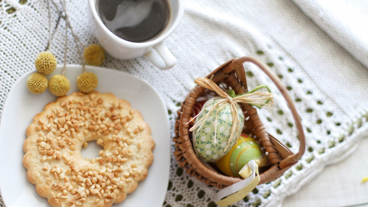 Coffee and a ring-shaped cookie beside a small basket of decorated Easter eggs on white lace