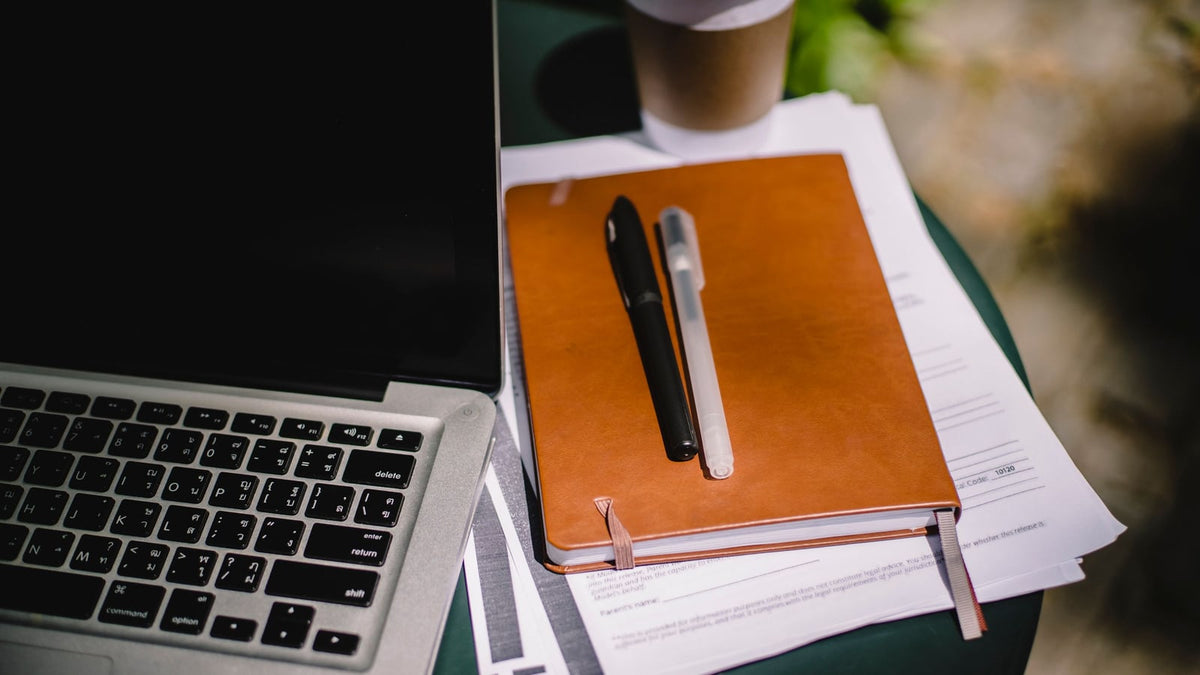A laptop, notebook, pens and takeaway coffee on a table for planning purchases