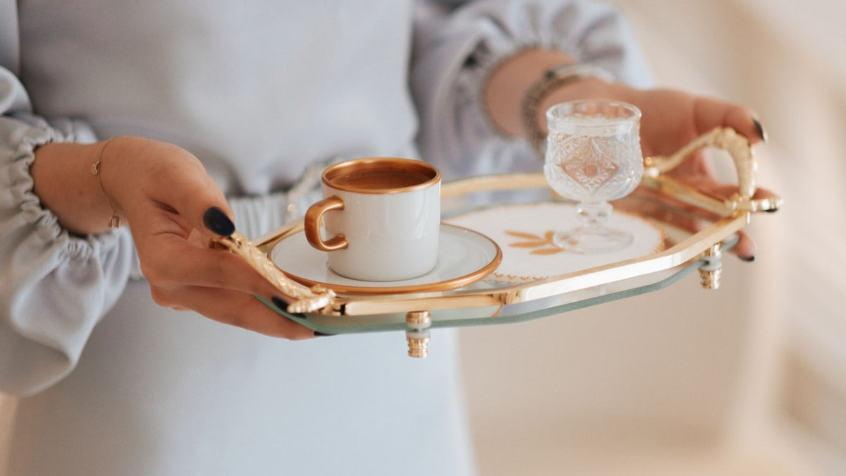 Hands carrying a gold-tone serving tray with a coffee cup and a glass of water