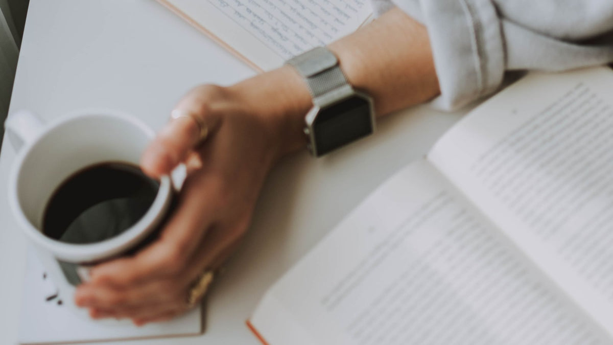 A hand holding a coffee cup beside an open handwritten notebook and book