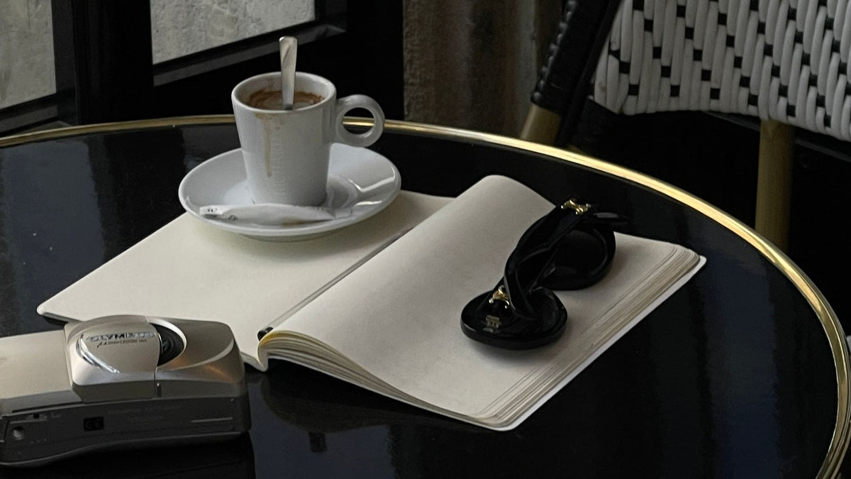 A cafe coffee cup with an open notebook, camera and sunglasses on a round table