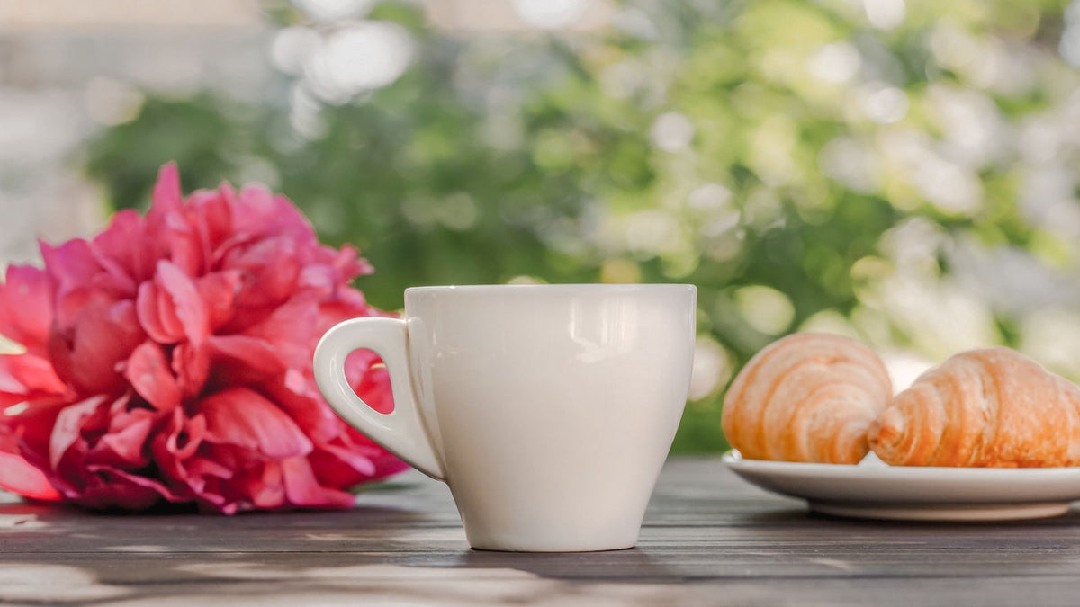 White coffee cup and croissants beside pink peonies on an outdoor wooden table